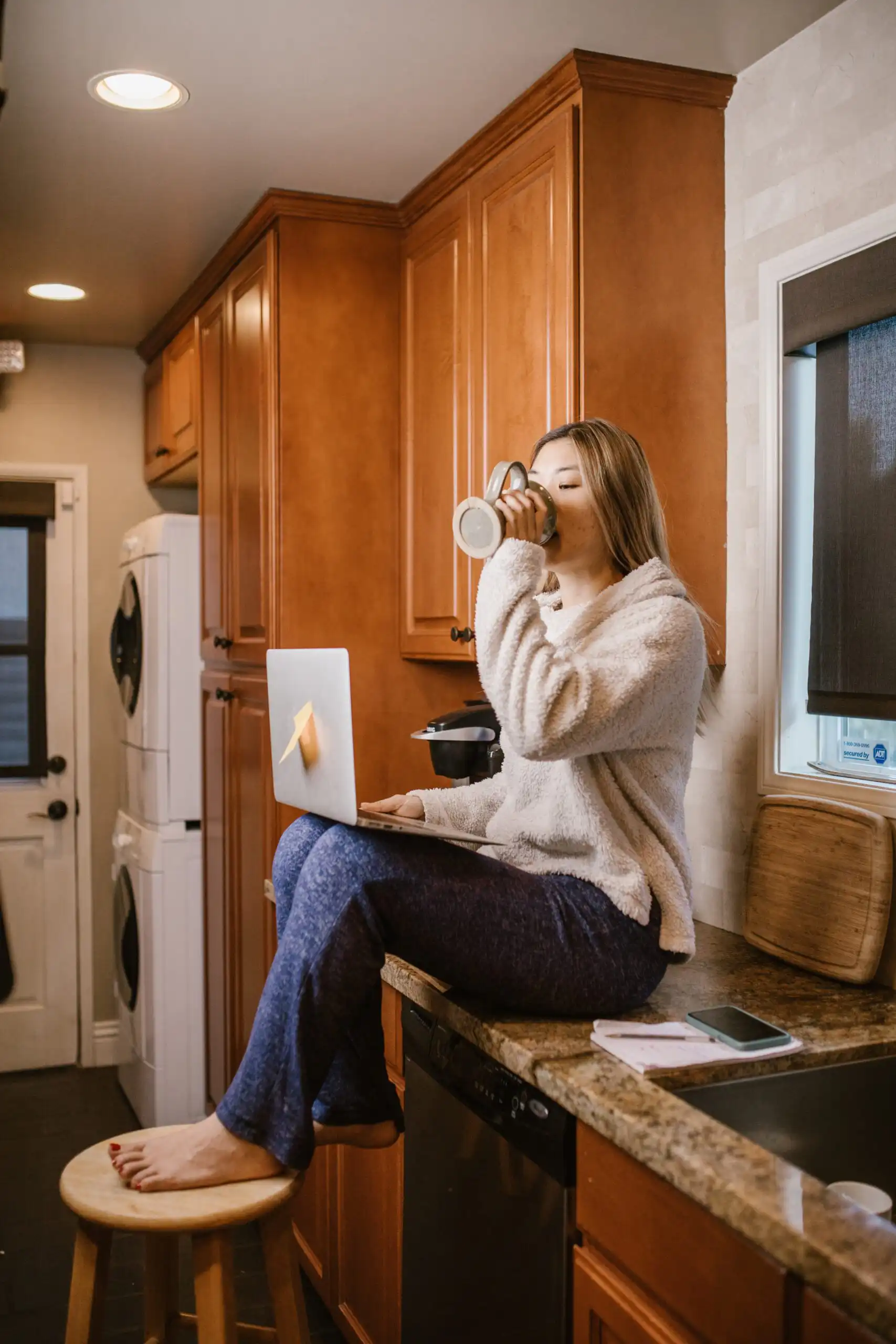 A young woman sits on her kitchen counter, drinking a beverage from a mug and working on the laptop propped on her thighs.